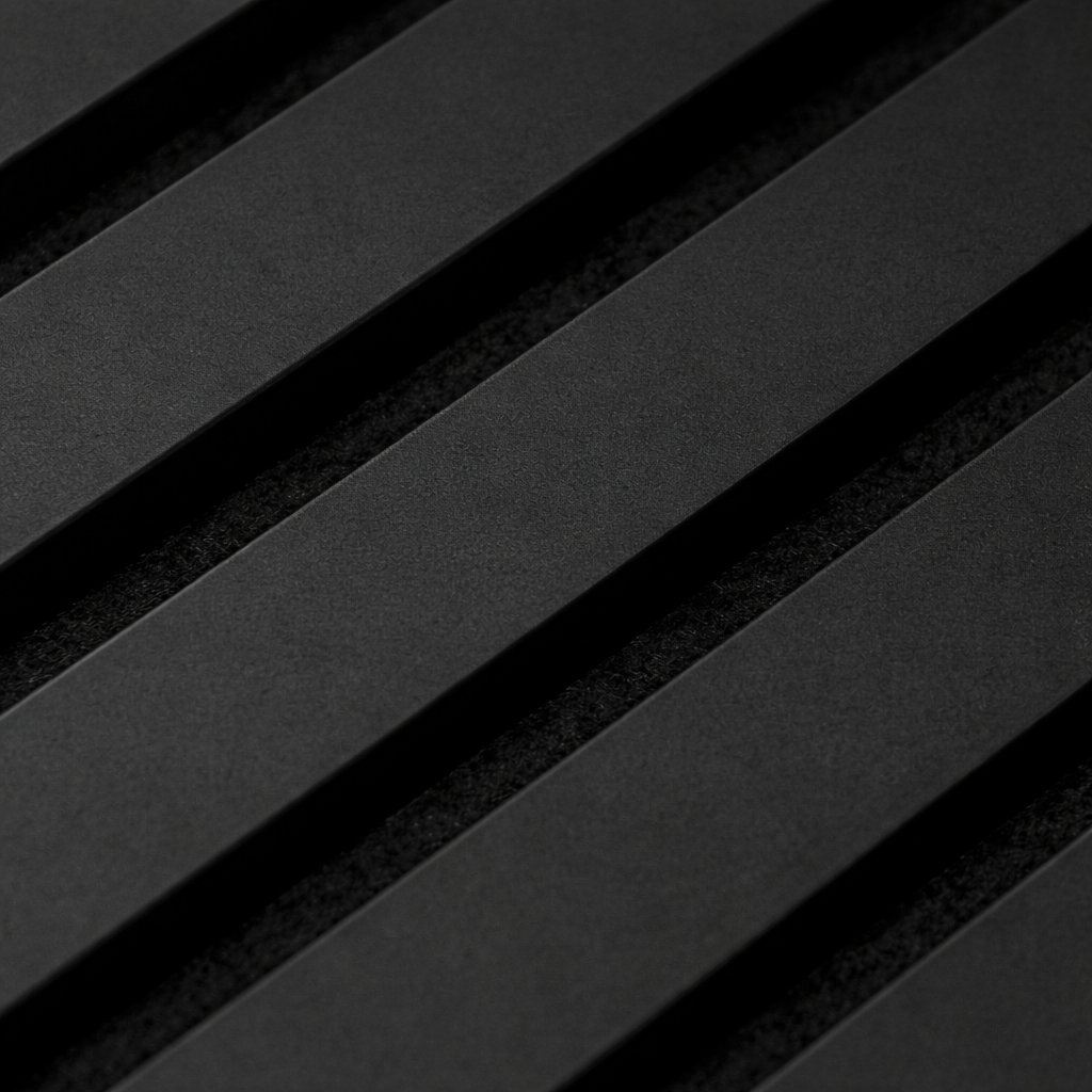 A close-up photograph of a charcoal black WoodUpp acoustic panel. The image reveals the textured surface of the black wooden slats and the fibrous black felt backing in the grooves. The diagonal perspective emphasizes the depth and construction of this sound-absorbing wall panel.