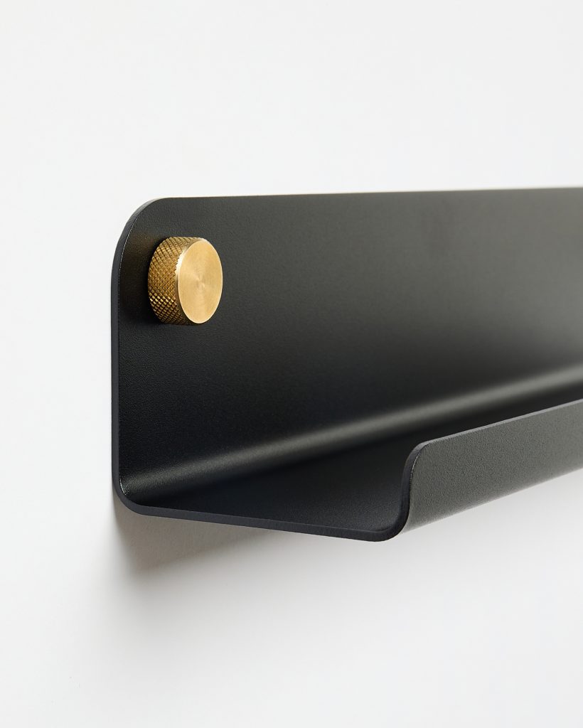 WoodUpp black wall-mounted shelf in matte powder-coated steel with a curved front lip, shown fixed to the wall by a large knurled brass mounting knob. Minimalist floating-design shelf with smooth rounded edges and an integrated pocket for holding mail, phones, or small accessories.