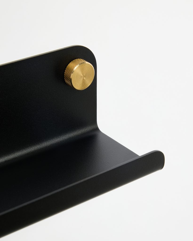 WoodUpp black powder-coated metal wall shelf close-up showing a minimalist L-shaped shelf with rounded front lip and textured finish, secured to the backplate by a knurled brass mounting knob. The image highlights material contrast and the WoodUpp shelf’s sleek, industrial design.