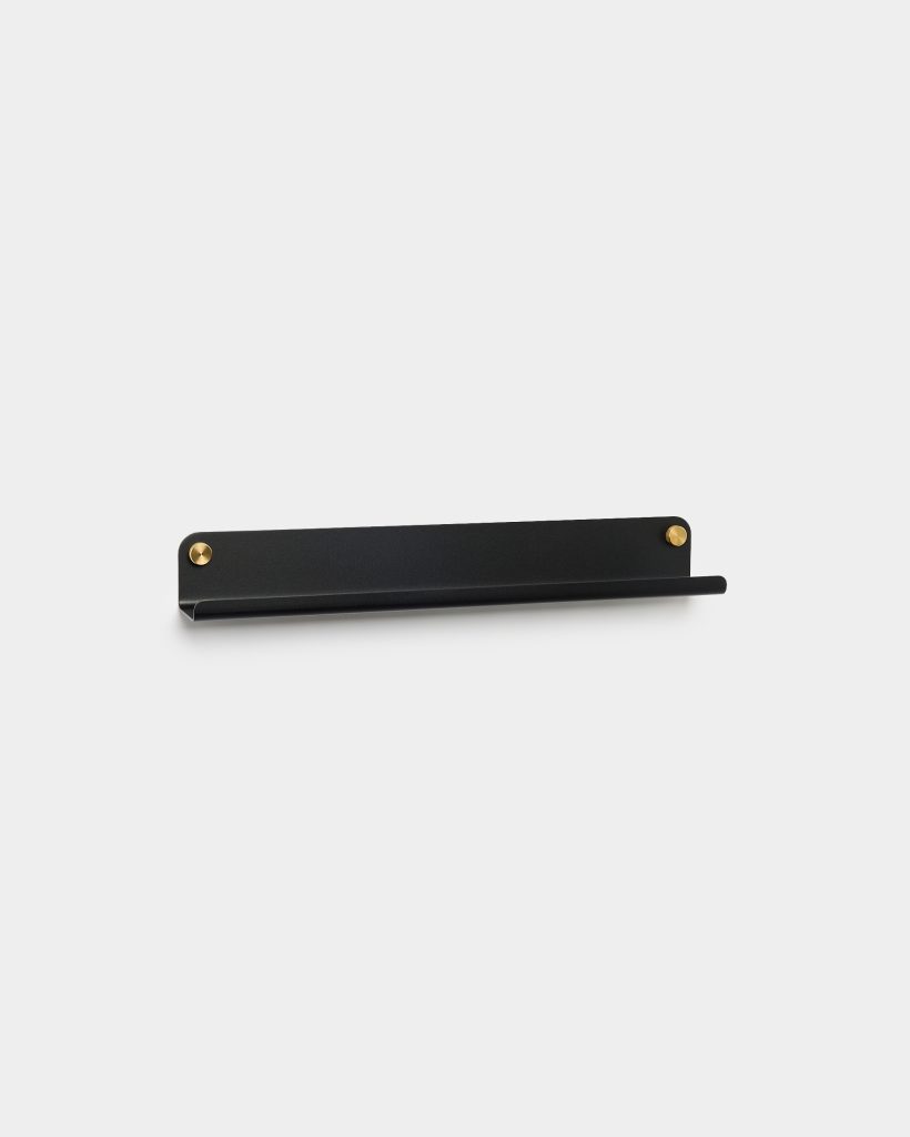 WoodUpp black floating shelf with integrated cylindrical front rail and slim lip, shown mounted on a white wall. Matte black metal shelf secured by two round brass fixings at each end, minimalist design ideal for small items, frames, decor, or toiletries.