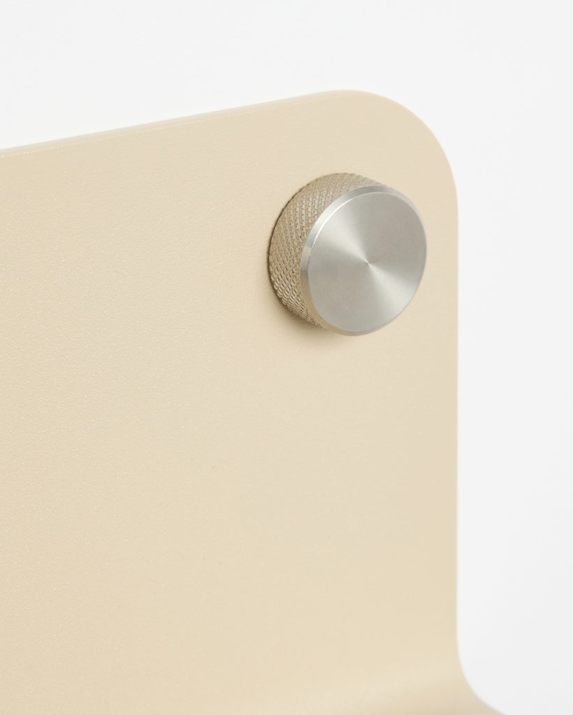 Close-up of a WoodUpp beige powder-coated metal panel with a rounded corner, showing a mounted knurled metal standoff/knob — a textured gold-toned cylinder topped with a brushed silver circular cap, used as a decorative mounting or adjustment control on the WoodUpp product.