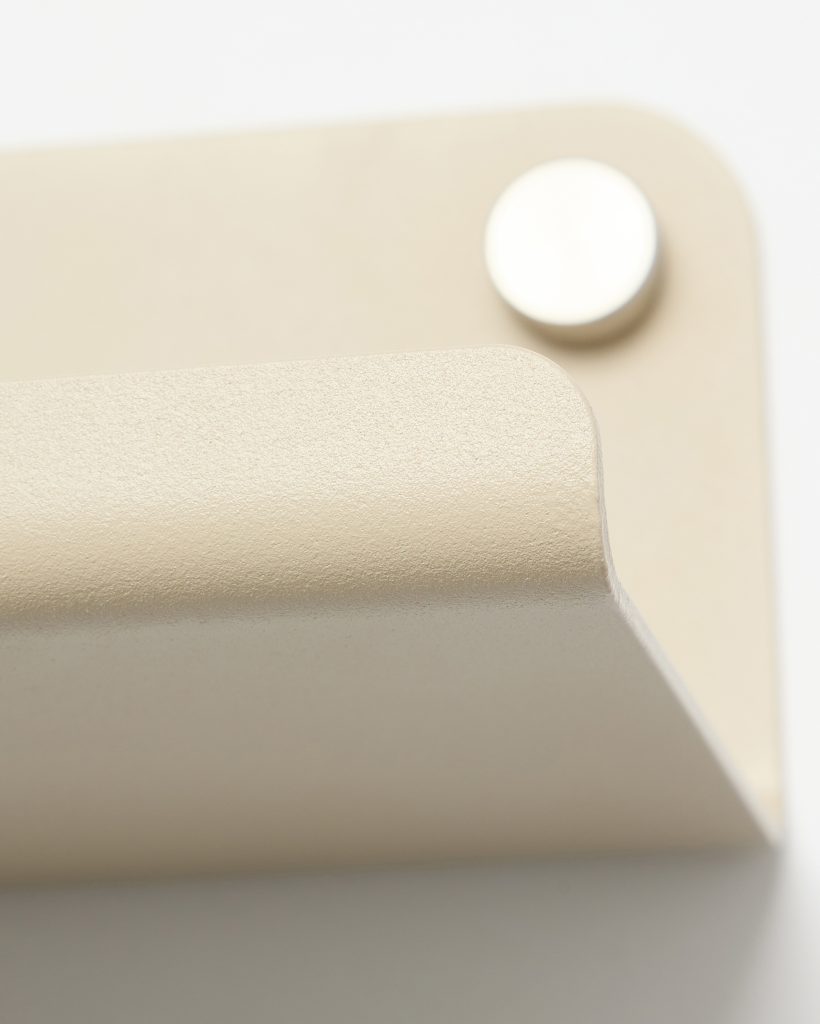 Close-up of WoodUpp's matte beige wall hook: a curved, textured metal lip mounted on a square backing plate, shown with a small circular silver mounting cap or magnet in the background. The image highlights the product’s smooth rounded edges, subtle grainy finish and minimalist design.