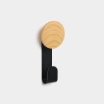 WoodUpp wall hook featuring a circular light oak wooden knob paired with a matte black metal S-shaped hook, mounted on a white wall. The minimalist WoodUpp coat hook shows visible wood grain, a slim vertical metal backplate and a rounded lower hook for hanging coats, bags or towels.