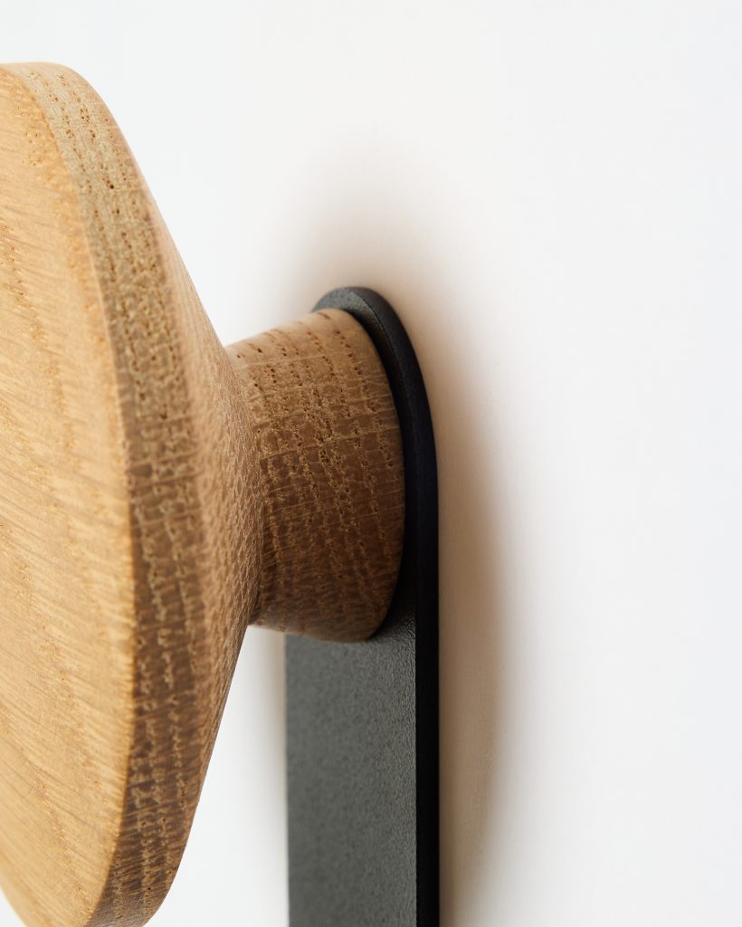 Close-up of a WoodUpp oak wall hook: a round, handcrafted oak peg with visible grain and tapered neck mounted on a matte black metal wall strip. The photo shows the WoodUpp wooden knob and its slim black bracket fixed to a white wall, highlighting texture and finish.