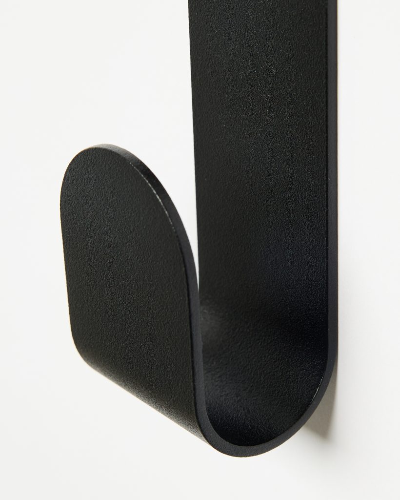 Close-up of a WoodUpp black wall hook: a single matte, textured J-shaped metal hook with a flat vertical backplate and a rounded forward lip, curved into a smooth U form. Mounted on a white wall, the minimalist hook emphasizes clean lines and sturdy, modern design.