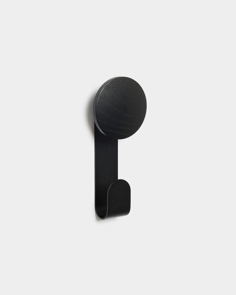 WoodUpp wall hook: single black wall-mounted coat hook featuring a round wooden knob with visible wood grain and a matte black curved metal strap forming the lower hook. Minimal, contemporary design shown against a light background, suitable for coats, towels, or bags.