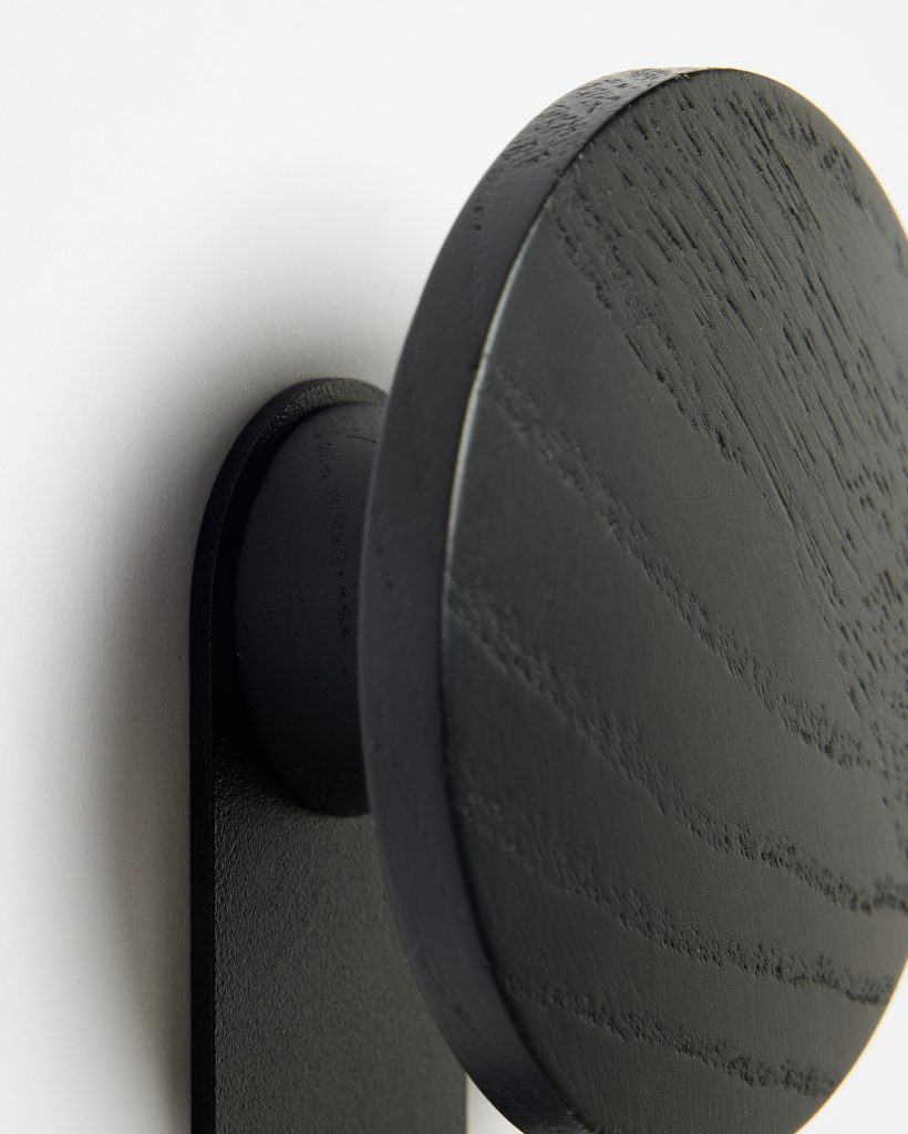 Close-up of a WoodUpp black wooden wall hook: a matte black circular disc with visible wood grain mounted on a slim rectangular backplate, connected by a rounded peg, shown against a white wall highlighting texture, finish and minimalist Scandinavian aesthetic