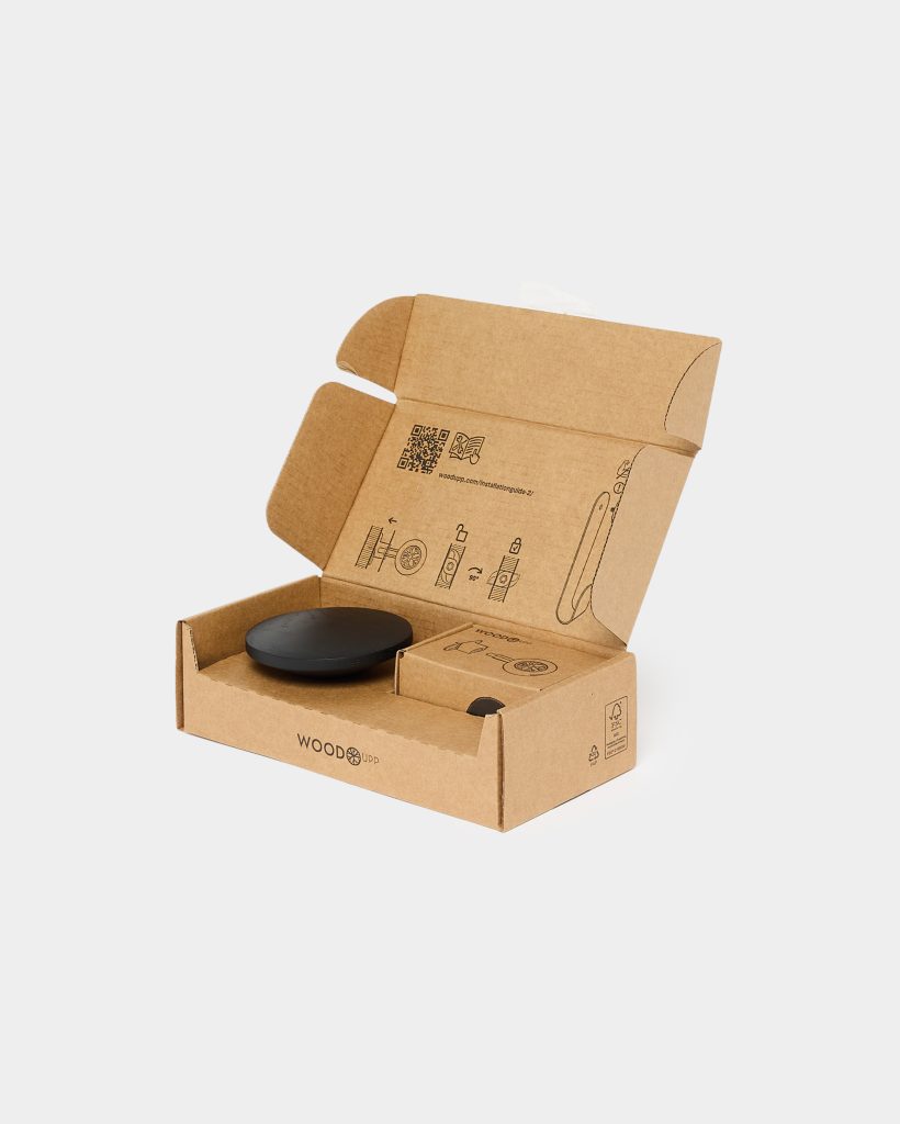 Open WoodUpp cardboard box revealing a matte black round puck-shaped smartphone mount resting in a molded insert, a small companion accessory box, and printed installation diagrams on the lid; the brown recyclable packaging bears the WoodUpp logo and simple line-art assembly instructions.