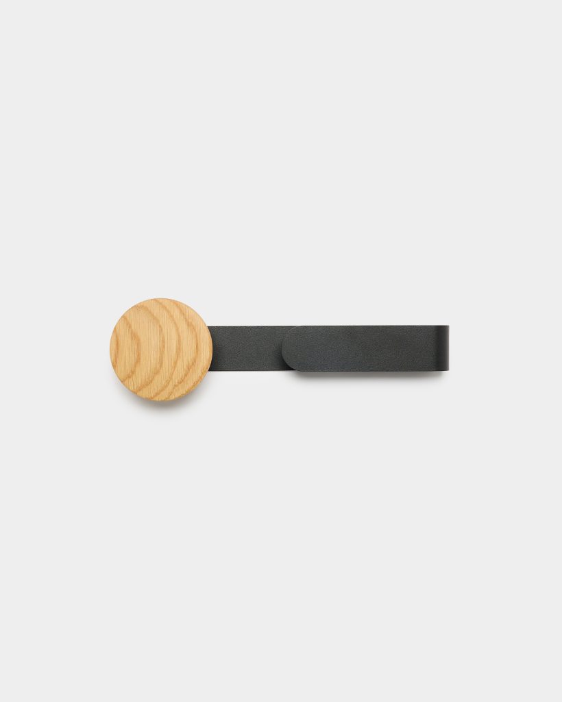 WoodUpp wall hook featuring a round natural oak wooden knob with visible grain attached to a matte black metal strap forming a slim horizontal loop. The minimalist wooden peg and black metal hanger are shown against a neutral white background, highlighting texture and simple Scandinavian design.