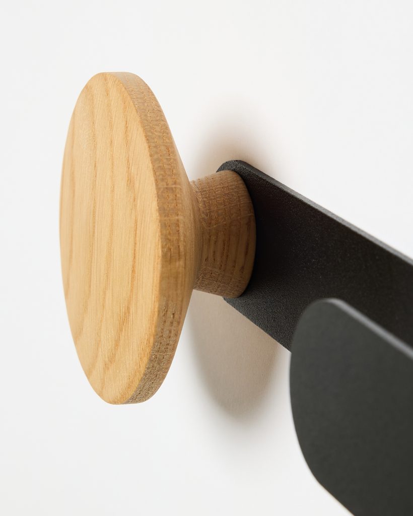 Close-up of a WoodUpp wall hook: a round oak knob with visible grain and turned cylindrical stem mounted on a matte black metal bracket, shown attached to a white wall — a minimal, Scandinavian-style wooden peg and metal wall plate from WoodUpp.