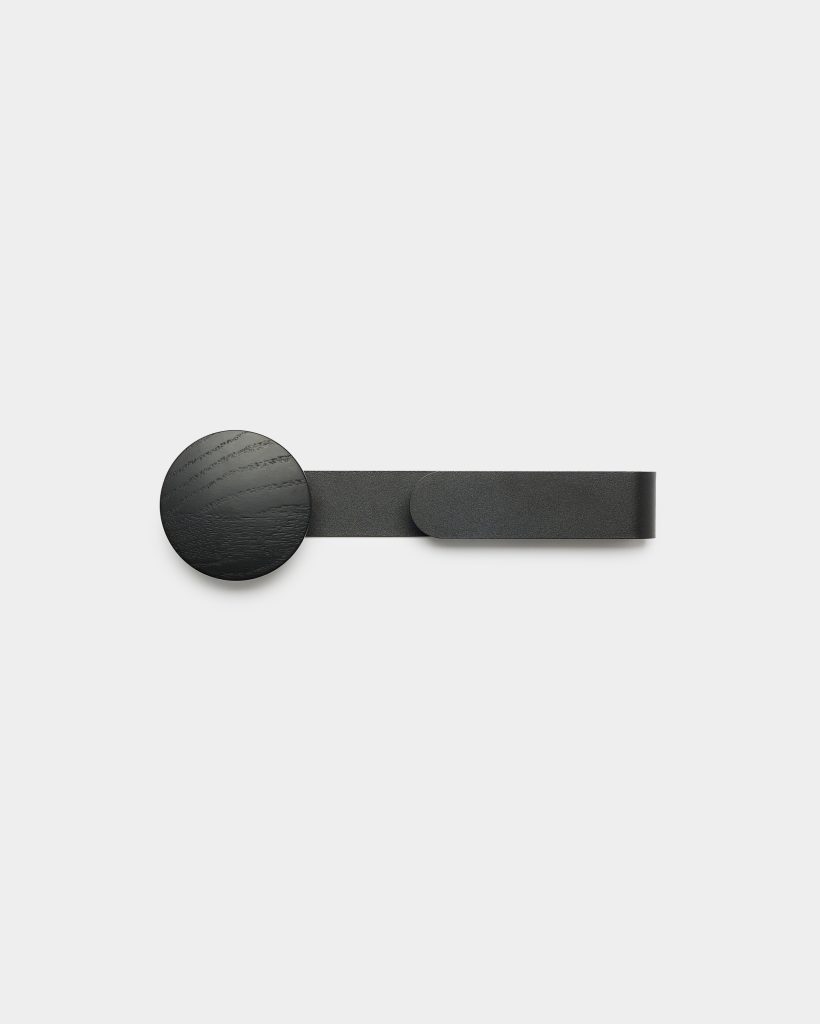 WoodUpp wall hook featuring a matte black round wooden knob with visible grain attached to a slim matte black metal strap and rounded end. The minimalist horizontal hook shows textured wood and metal finishes against a pale background, highlighting WoodUpp’s simple, modern hardware design.