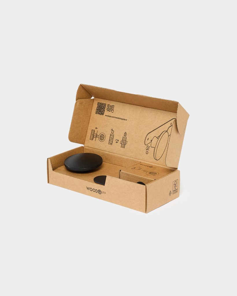 WoodUpp packaging open to show two matte black wooden wall hooks — one larger disc and one smaller disc — plus a small box of mounting hardware, all seated in a recycled cardboard box printed with simple assembly icons and the WoodUpp logo.