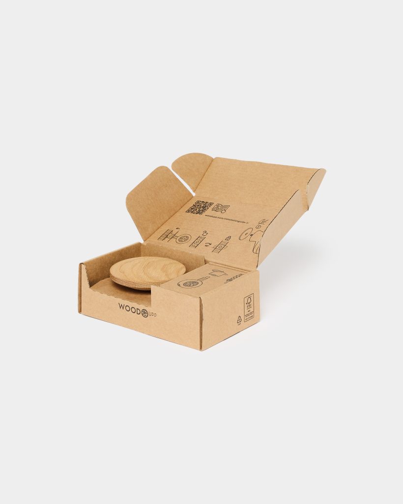 Open WoodUpp kraft cardboard packaging displays a stacked pair of round, natural oak wooden coasters nestled inside. The box features printed assembly icons, a QR code and the WoodUpp logo on the front, emphasizing eco-friendly recycled materials and minimalist, protective product presentation.