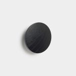 WoodUpp circular black wooden wall hook mounted on a white background, showing visible wood grain and a matte finish. The compact, disc-shaped coat hook projects slightly from the wall, casting a soft shadow — a minimalist WoodUpp accessory ideal for hanging coats, towels, or bags.