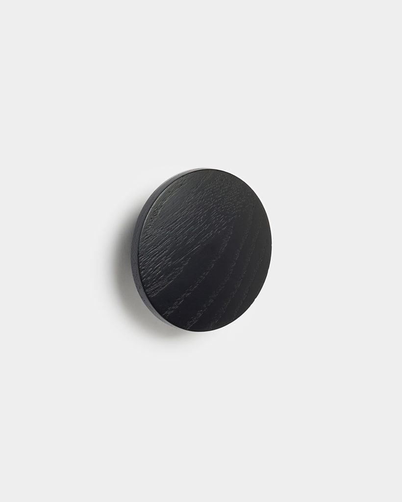 WoodUpp circular black wooden wall hook mounted on a white background, showing visible wood grain and a matte finish. The compact, disc-shaped coat hook projects slightly from the wall, casting a soft shadow — a minimalist WoodUpp accessory ideal for hanging coats, towels, or bags.
