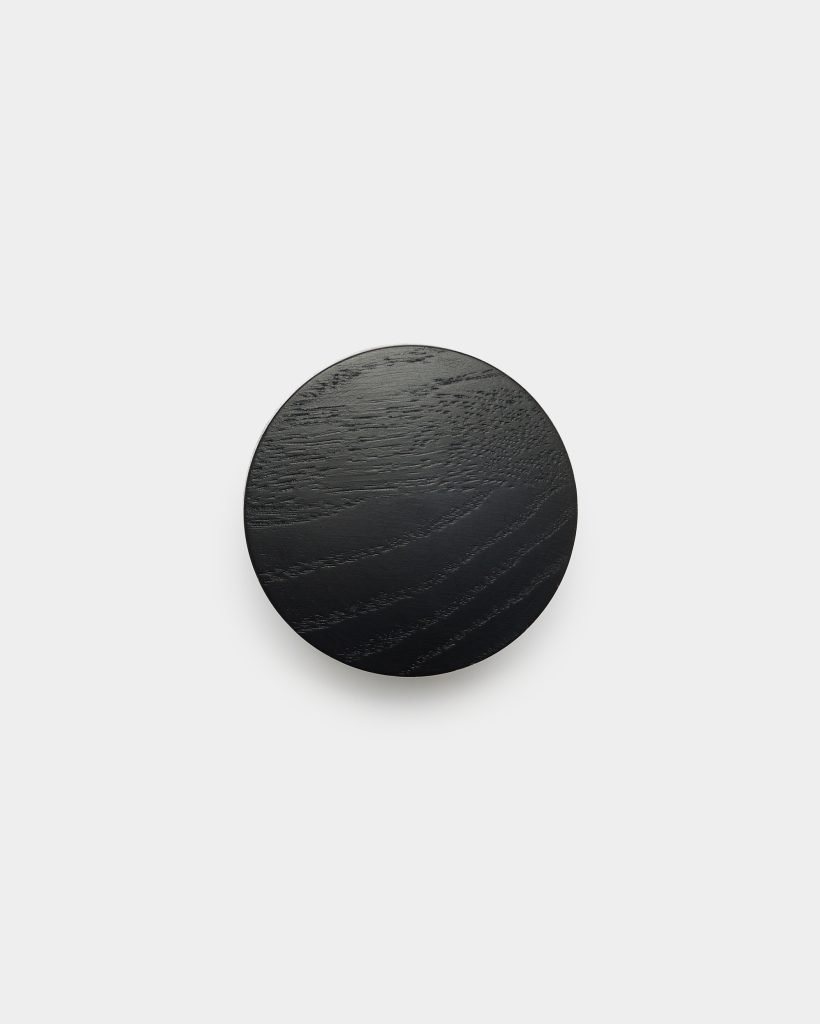 WoodUpp black circular wooden wall hook photographed against a white background. Smooth, matte-finished round knob shows visible oak grain, thin beveled edge and soft shadow below, suggesting a mounted minimal coat or towel hook. Simple, modern Scandinavian design with an elegant finish.