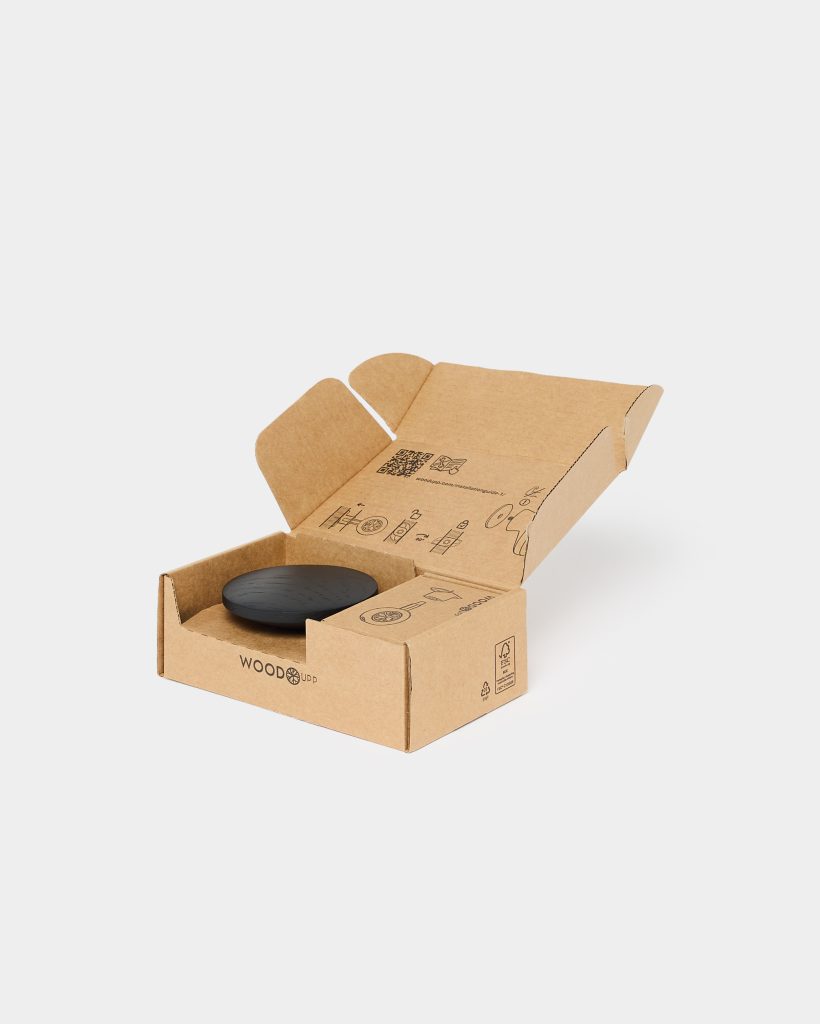 Open WoodUpp branded recycled cardboard box with printed assembly icons, containing a matte black round wooden disc and a compact rectangular accessory box nestled in a die-cut insert. Packaging shows the WoodUpp logo and recycling/FSC marks on the outer panels.