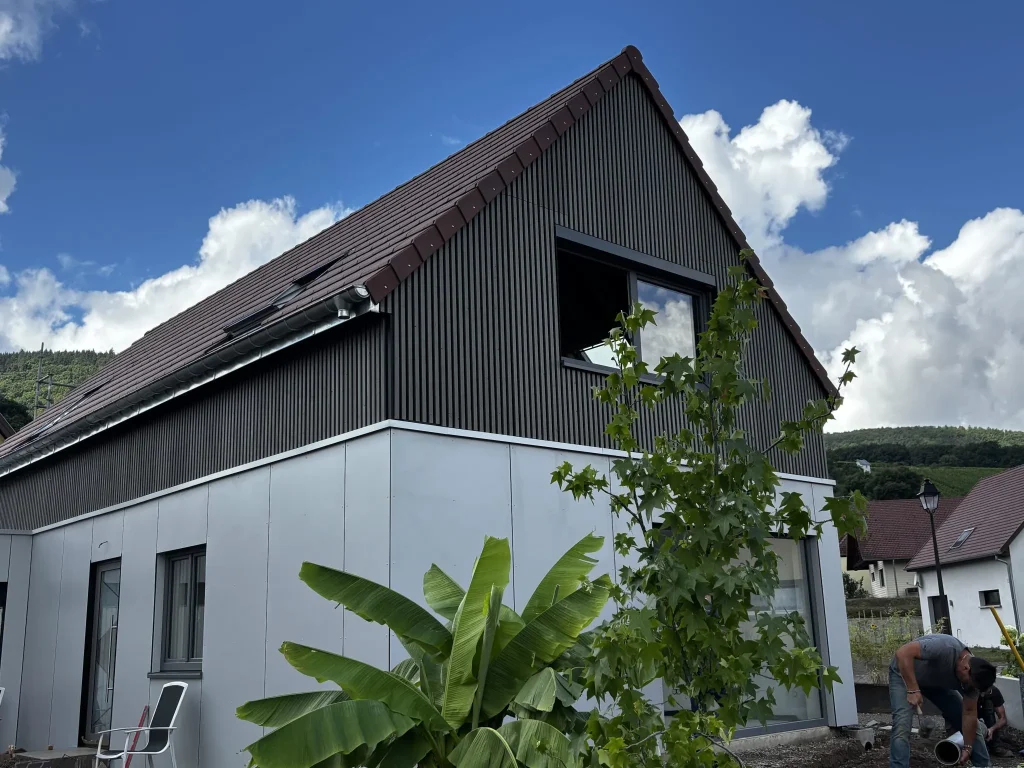 Modern two-story house clad with WoodUpp vertical dark timber slats on the upper gable and light grey WoodUpp external panels on the ground floor, a black-framed window, banana plant and young tree in foreground, and a worker kneeling at right installing exterior drainage.