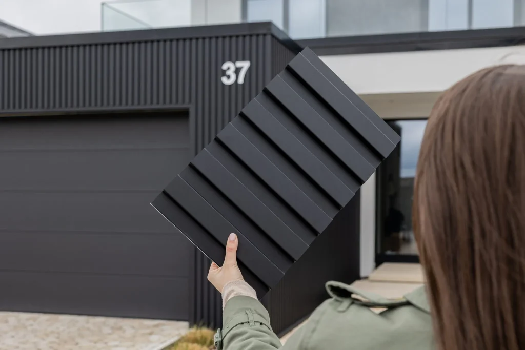 Person holds a WoodUpp black ribbed sample panel in front of a modern home clad in matching WoodUpp vertical slat exterior cladding; the handheld sample shows deep, evenly spaced horizontal grooves while the house features matte anthracite slats around the garage and entrance.
