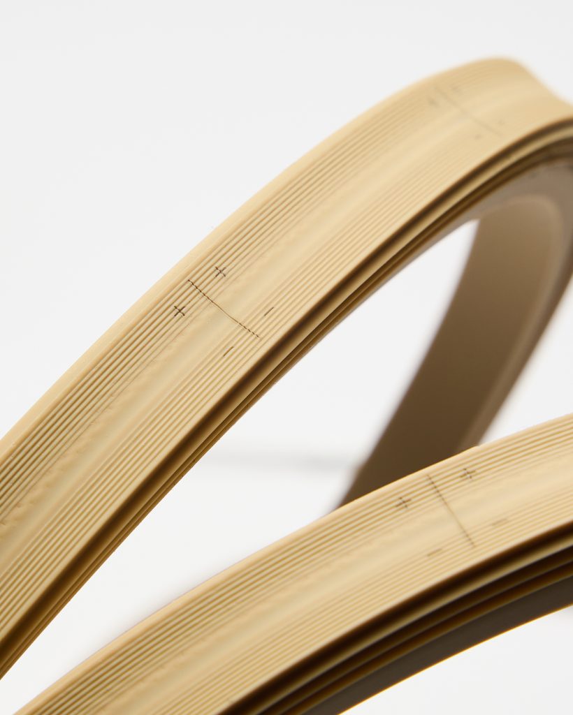 Close-up of two curved WoodUpp beige edge banding strips: flexible PVC edge trim with fine parallel ridges and printed alignment marks and measurement ticks, shown arched against a white background. The WoodUpp products display textured ribbing and guide markings for precise application on furniture edges.