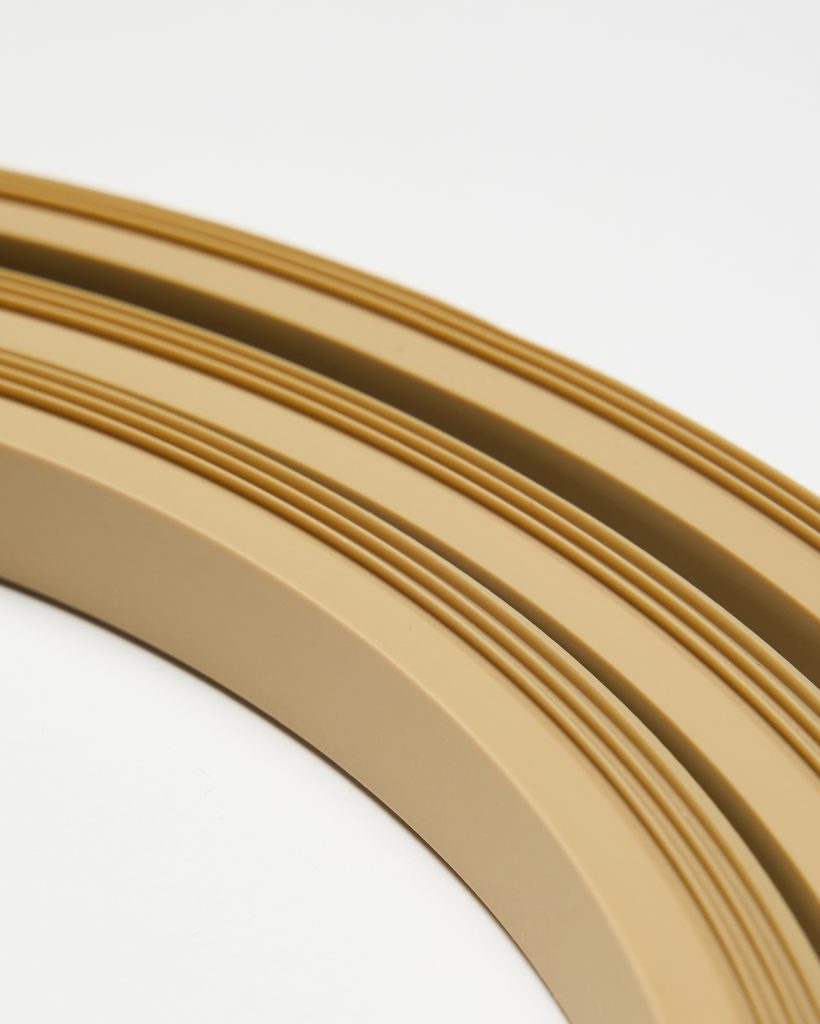 Close-up of WoodUpp beige curved trim strips: three stacked semicircular edging profiles with fine parallel grooves, smooth matte finish and shallow channels. The flexible polymer edging pieces appear uniform in colour and thickness, shown against a white background and photographed at an angled macro perspective.