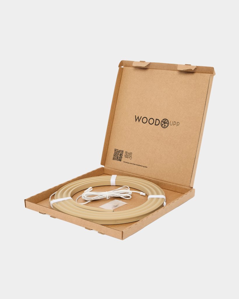 Open WoodUpp cardboard box displaying the WoodUpp logo, containing a coiled light oak-colored wooden LED profile strip, bundled white power cable, small connector and adhesive mounting pads. The minimalist packaging showcases the flexible wood-toned edging/LED trim ready for installation immediately.