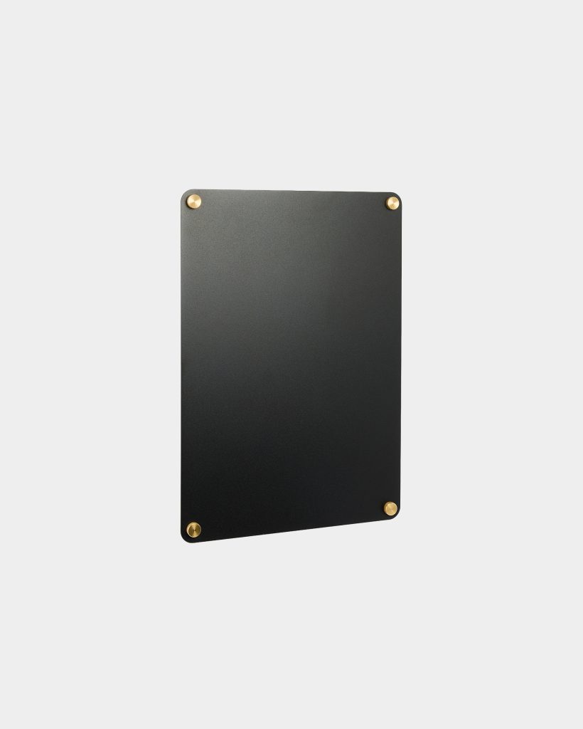 WoodUpp black rectangular wall board with a smooth matte finish shown against a pale background, fastened at each corner by four round brass mounting studs. The minimalist memo board appears thin and magnetic, ideal for notes or photos, showcasing WoodUpp’s modern, industrial design aesthetic.