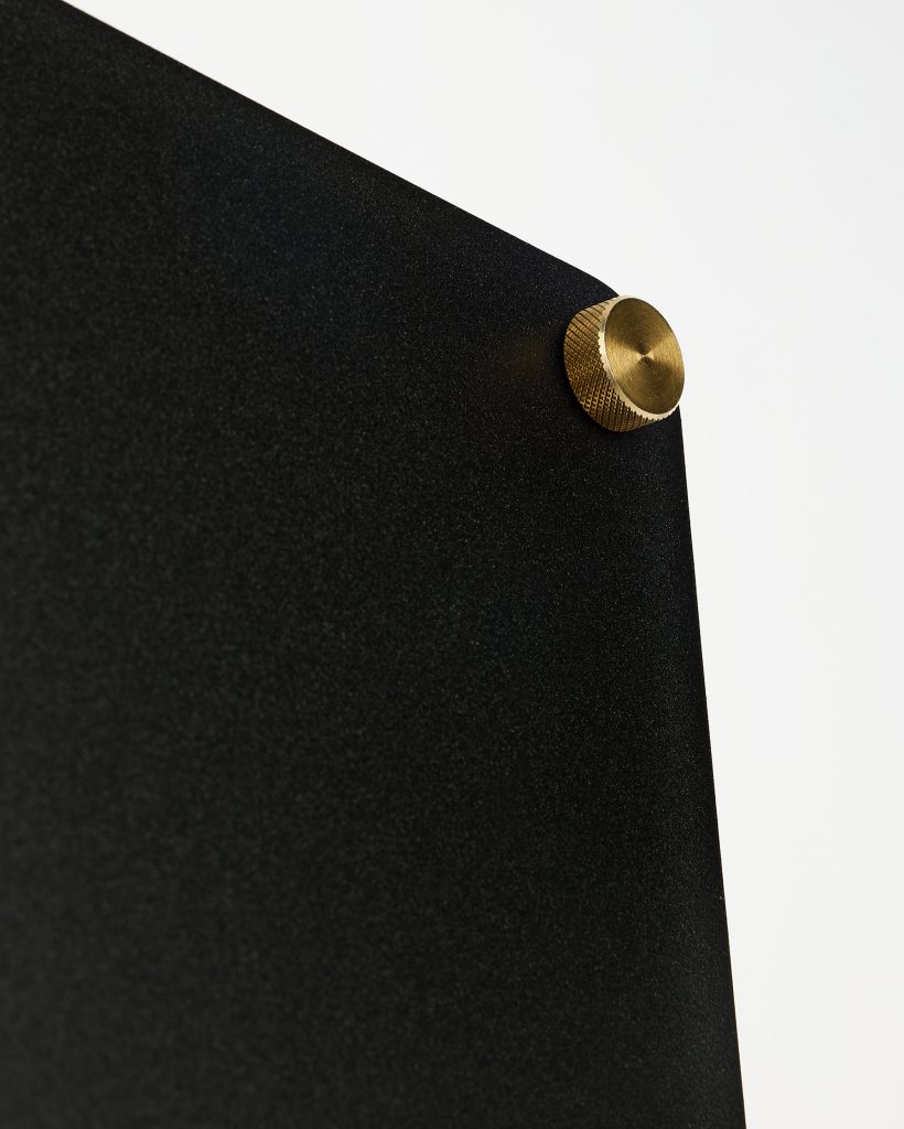 WoodUpp black textured display panel held by a single brass knurled mounting screw/standoff. The close-up shows the matte black panel surface and the gold-tone metal knob with fine knurled grip, highlighting WoodUpp’s minimalist wall-mounted panel and brass fixing hardware detail.