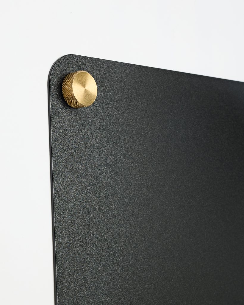 WoodUpp black textured magnetic wall board with rounded corner mounted on a white wall, secured at the top-left by a brass knurled mounting screw/standoff. The gold-toned circular brass pin has a smooth face and cross-hatched edge, contrasting with the matte black board surface.