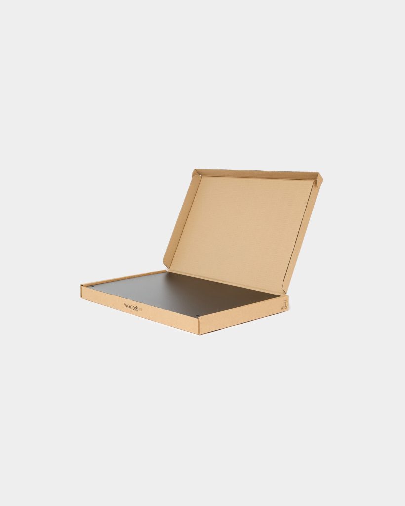 Open WoodUpp cardboard mailer on a white background, containing a matte black rectangular panel from WoodUpp. The slim panel sits flat inside the box, showing rounded corners and four small corner holes; the WoodUpp logo is printed on the box edge, emphasizing branded packaging.