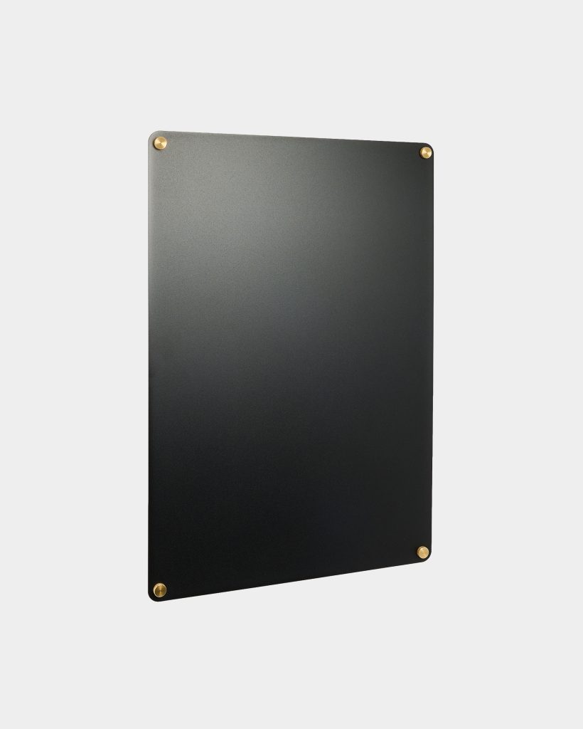 WoodUpp black magnetic board shown against a pale grey background — a smooth matte rectangular panel slightly angled, held at its four rounded corners by small round brass standoffs; minimalist vertical display board with subtly rounded edges and a sleek, contemporary finish.