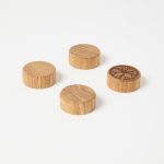 WoodUpp product photo shows four small round wooden plugs: light-brown, finely grained discs with smooth sides. Three have plain tops while one features a dark, laser-engraved floral starburst pattern. The WoodUpp round wooden plugs are displayed on a clean white background.