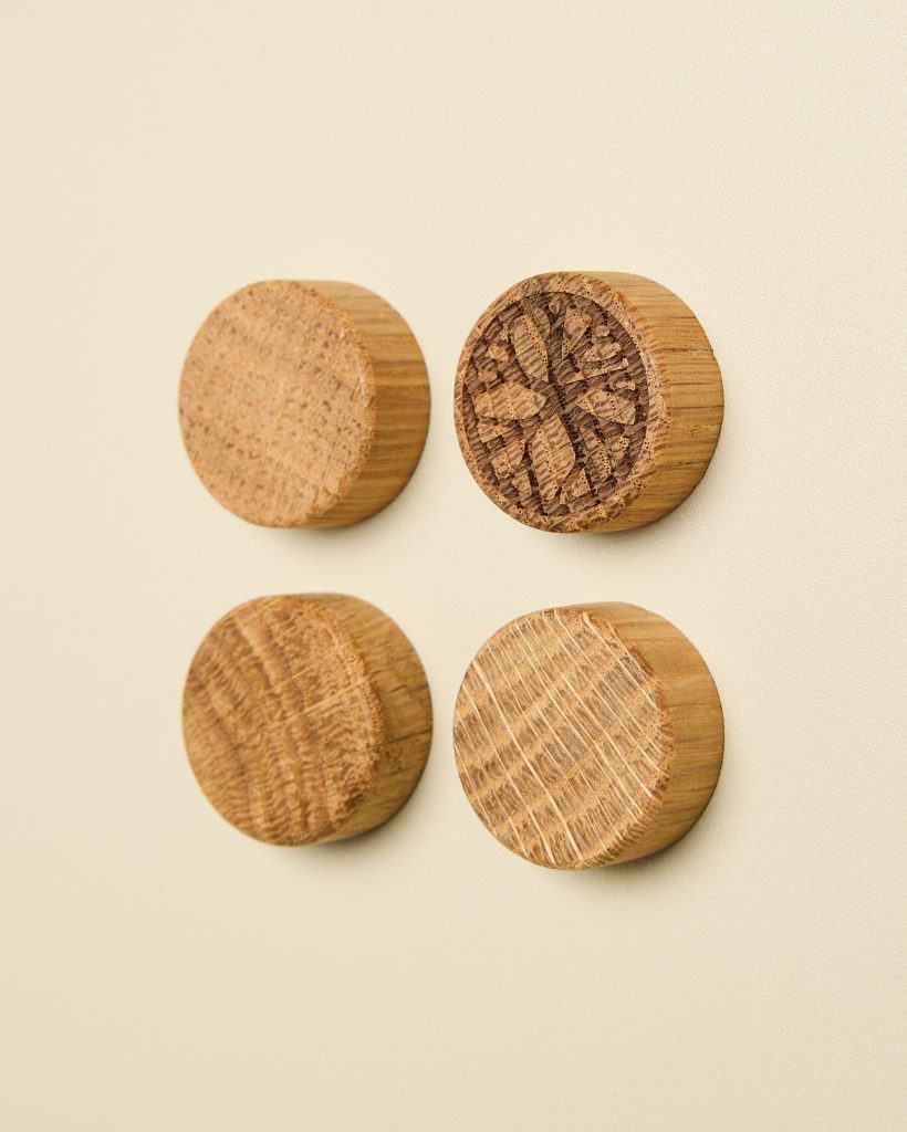 Four WoodUpp round oak magnets arranged in a 2x2 grid on a pale cream surface. Each cylindrical wooden magnet shows natural grain; the top-right piece features a carved floral motif. Smooth, light-stained finish highlights texture and concentric growth rings across the compact wooden discs.