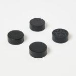 Four WoodUpp black wooden discs sit on a white background — small round, low-profile tokens with visible wood grain; three plain matte discs and one engraved with a floral starburst motif, casting soft shadows, arranged roughly in an elegant loose square.