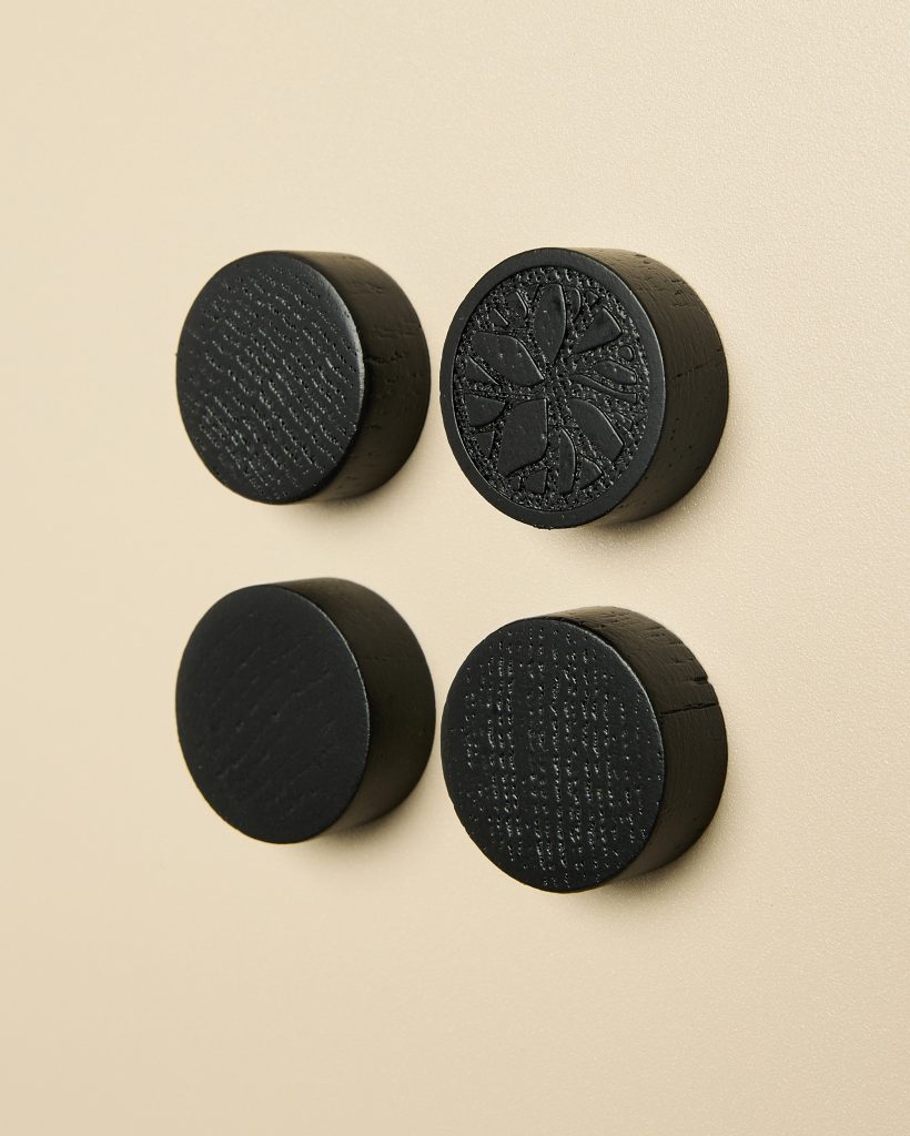 Four WoodUpp round black wooden magnets arranged in a square on a pale beige surface; three are smooth with visible wood grain, while the top-right magnet features an engraved floral mandala pattern, all cylindrical disk-shaped pieces with matte, textured finish.
