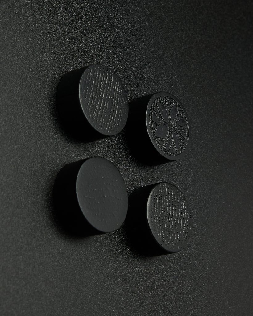 WoodUpp: four matte black wooden discs mounted on a textured black surface, arranged in a two-by-two grid. Three discs show subtle wood-grain texture; the fourth has a carved radial, tree-like motif. These WoodUpp pieces resemble magnetic wood studs or decorative wall plugs.