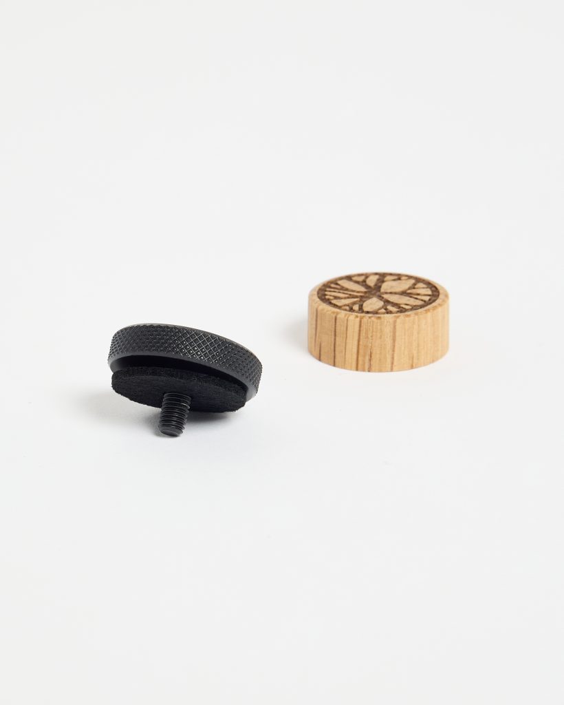 WoodUpp image showing two products: a small round oak wooden cap with laser-etched floral design, and a black metal threaded mounting knob with knurled edge and felt pad. Both items sit on a white background, illustrating WoodUpp’s decorative wooden caps and metal mounting adapters.