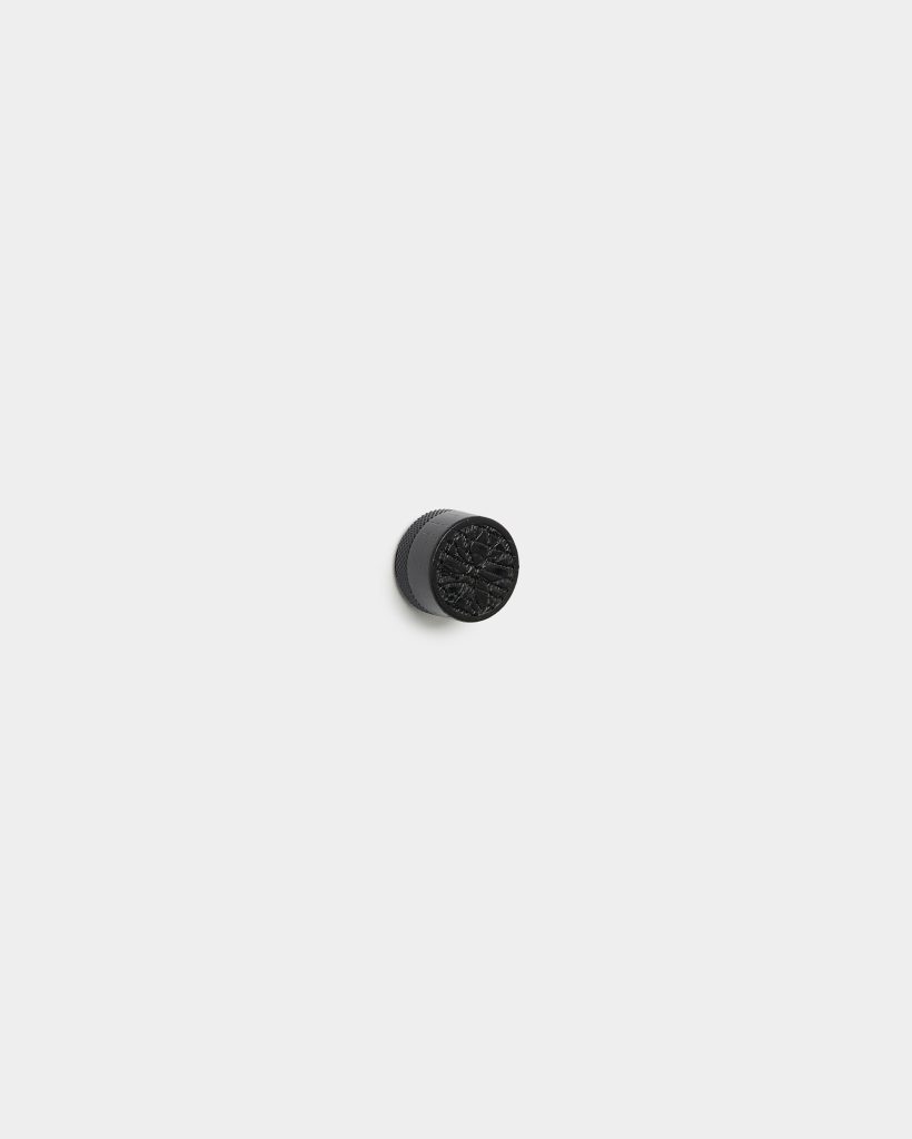 WoodUpp black round cabinet knob centered on a white background. The small metal knob features a knurled cylindrical side and a decorative cracked radial pattern on its top face, showing subtle matte finish and a shadow beneath. Simple product shot highlighting texture and shape.