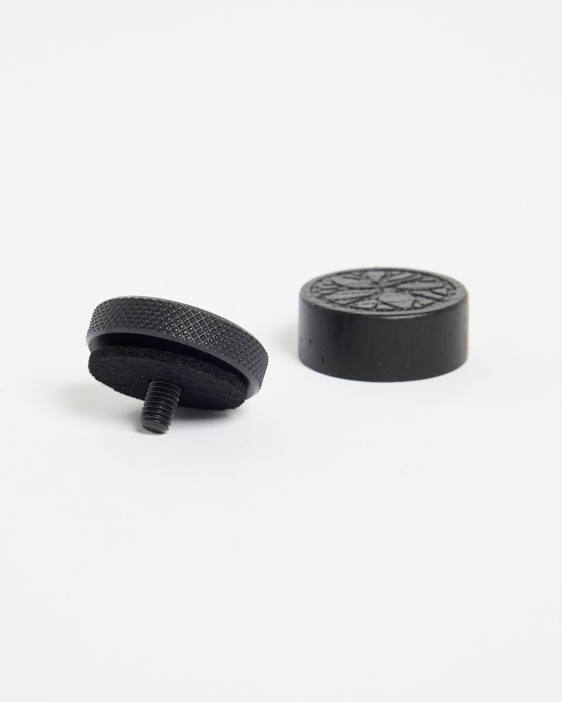 WoodUpp black furniture glides pictured: one threaded, knurled metal glide with adhesive felt pad underneath and exposed screw threaded stud; beside it a matching round solid rubber foot protector with decorative starburst pattern on top, designed to protect floors and stabilise wooden furniture.