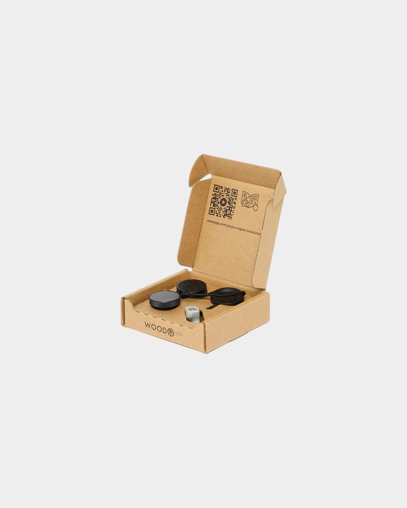 Open WoodUpp-branded cardboard box showing a poster mounting kit: two black textured round magnetic discs with attached fabric straps, a small silver metal mounting magnet, and plastic spacers, plus a printed QR code and installation icon on the inside lid. Clean white background.