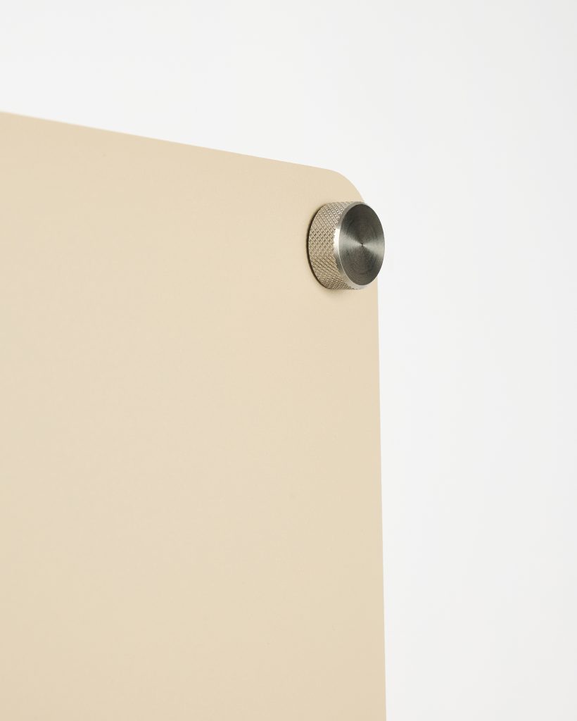 Close-up of a WoodUpp beige coated panel with rounded corner, mounted to a white background by a brushed stainless-steel knurled standoff; the WoodUpp board has a smooth matte finish while the metal standoff shows a circular brushed face and textured edge.