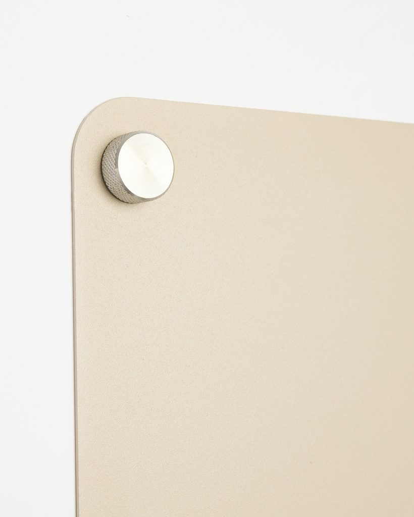 WoodUpp beige metallic wall panel with rounded corner mounted on a white wall, secured by a knurled silver aluminium standoff. The close-up shows the panel's fine textured finish and the cylindrical, ridged WoodUpp mounting knob holding the rectangular panel flush to the wall.