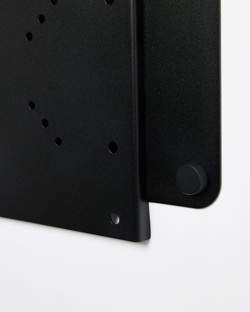 Close-up of a WoodUpp matte black powder-coated metal wall panel and matching accessory: a perforated mounting board with multiple circular holes and a textured round black knob/peg fixed to the panel’s edge, highlighting precise edges and a minimalist industrial finish for modular wall storage.