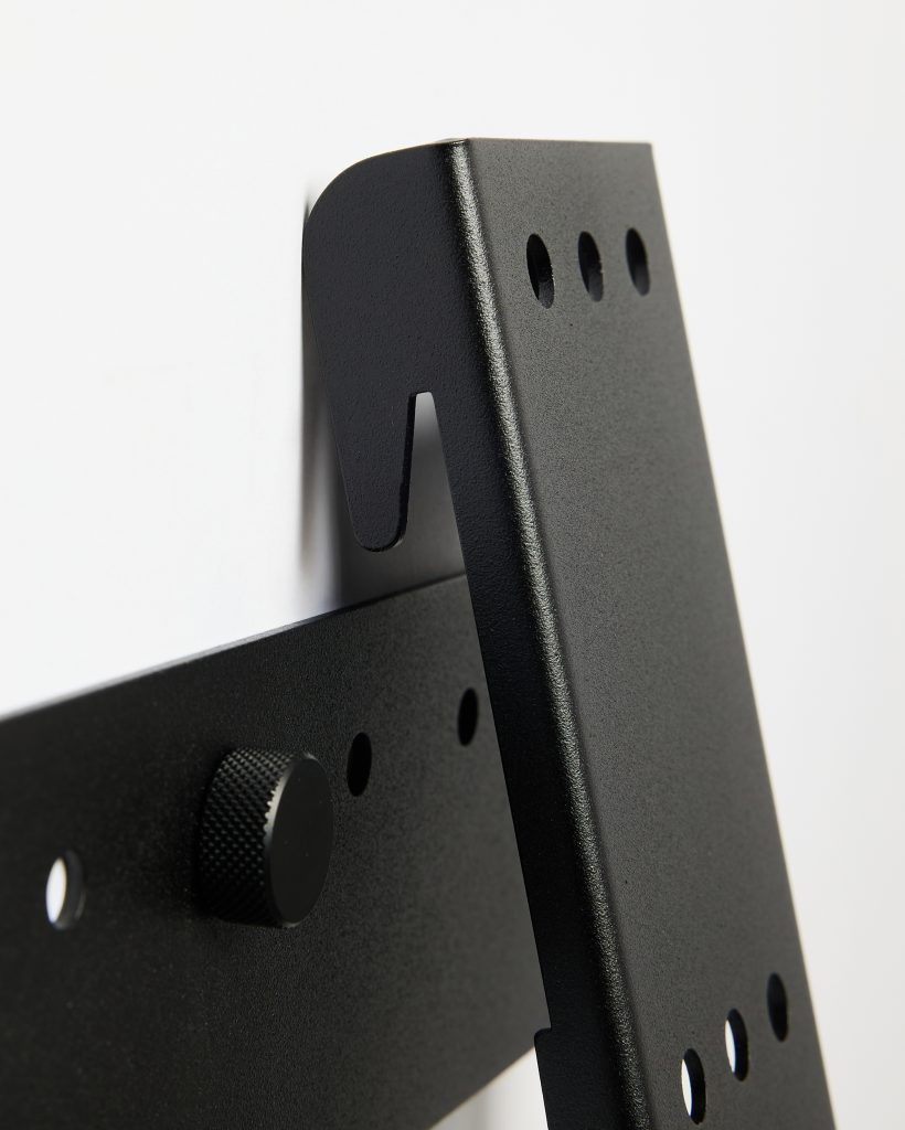 Close-up of WoodUpp black powder-coated steel wall-mount bracket, showing a slotted hanging hook, three circular VESA-style holes and a textured knurled adjustment knob. The heavy-duty WoodUpp bracket’s matte finish, rounded edges and mounting holes are visible against a plain white background.