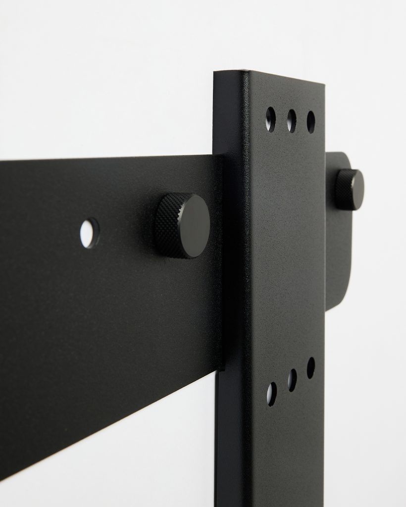 Close-up of WoodUpp sliding door hardware: matte black powder-coated steel vertical wall bracket and horizontal strap, showing three pre-drilled holes and two knurled round adjustment knobs/bolts. The detailed photo highlights screw holes, textured finish and sturdy bracket connection used in WoodUpp barn-door kits.