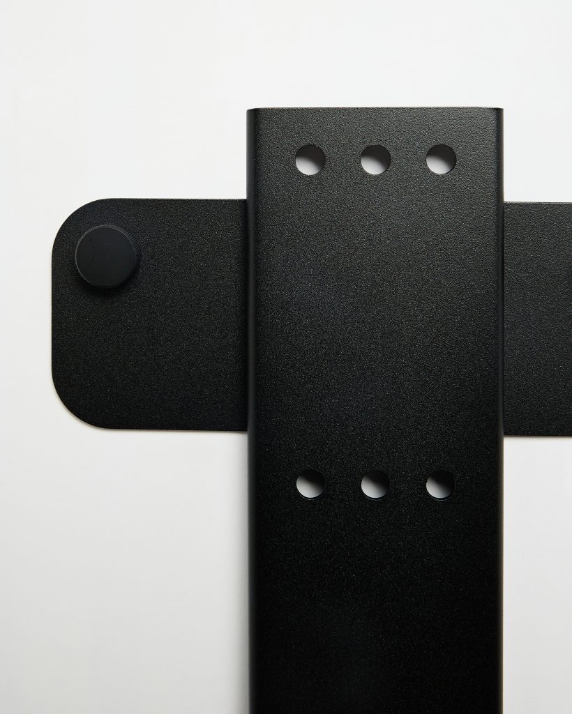 WoodUpp close-up of two intersecting matte black powder-coated metal mounting plates: a vertical, low-profile plate with three precision-drilled circular holes and a horizontal rounded plate with a textured rubber knob/stop. Sleek, minimal industrial hardware photographed against a clean white background.