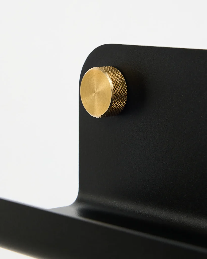Close-up of a WoodUpp matte black metal bracket with rounded corner and curved lower support, fitted with a circular knurled brass knob. The WoodUpp brass piece shows a textured grip around a smooth brushed face, contrasting with the flat, powder-coated black surface of the bracket.