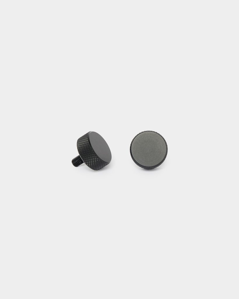 White background with two WoodUpp black knurled metal knobs laid side by side: one shows a short threaded stud and textured grip, the other displays a flat felt-padded base. Both are matte black, compact circular mounting knobs designed for furniture or accessory attachment.