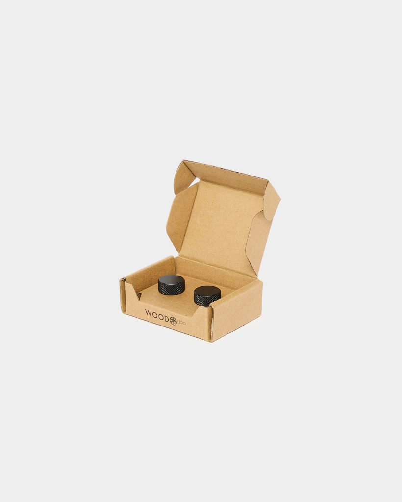 Open cardboard box branded WoodUpp contains two small black textured metal knob caps seated inside protective cardboard inserts. The compact kraft box is open, showing the pair of round, knurled-surface WoodUpp accessory pieces neatly fitted for shipping or display against a plain white background.