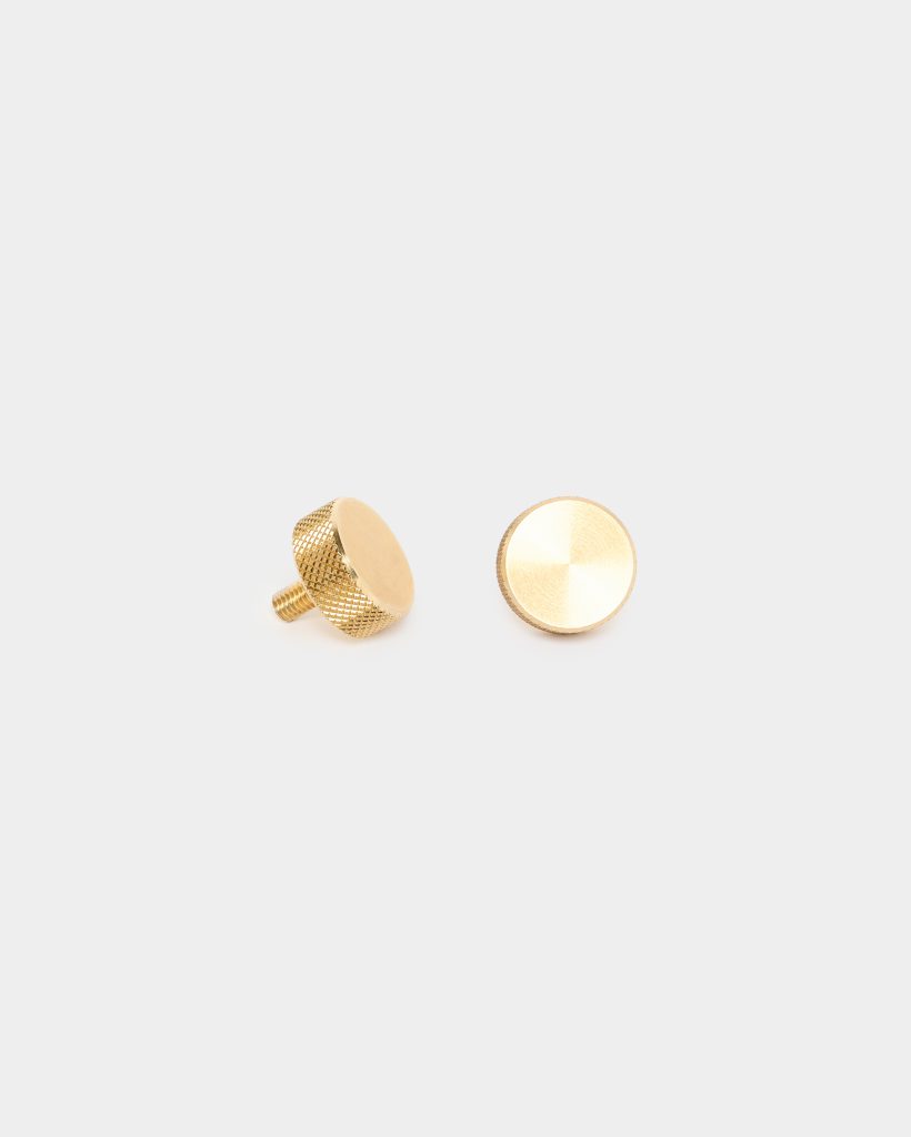 WoodUpp brass replacement knobs: two small round brass control knobs photographed on a white background. One displays a knurled textured side and exposed threaded stud, the other shows a smooth, brushed circular top. Both are matte gold-tone, compact hardware pieces from WoodUpp.